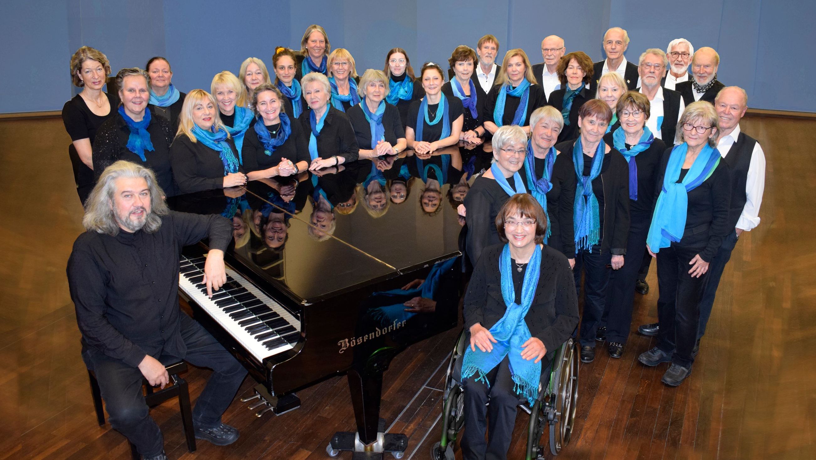 Foto des GofenbergChores in schwarzer Kleidung mit blauen Schals. Der Chor steht an einem Flügel, der Chorleiter sitzt am Flügel.
